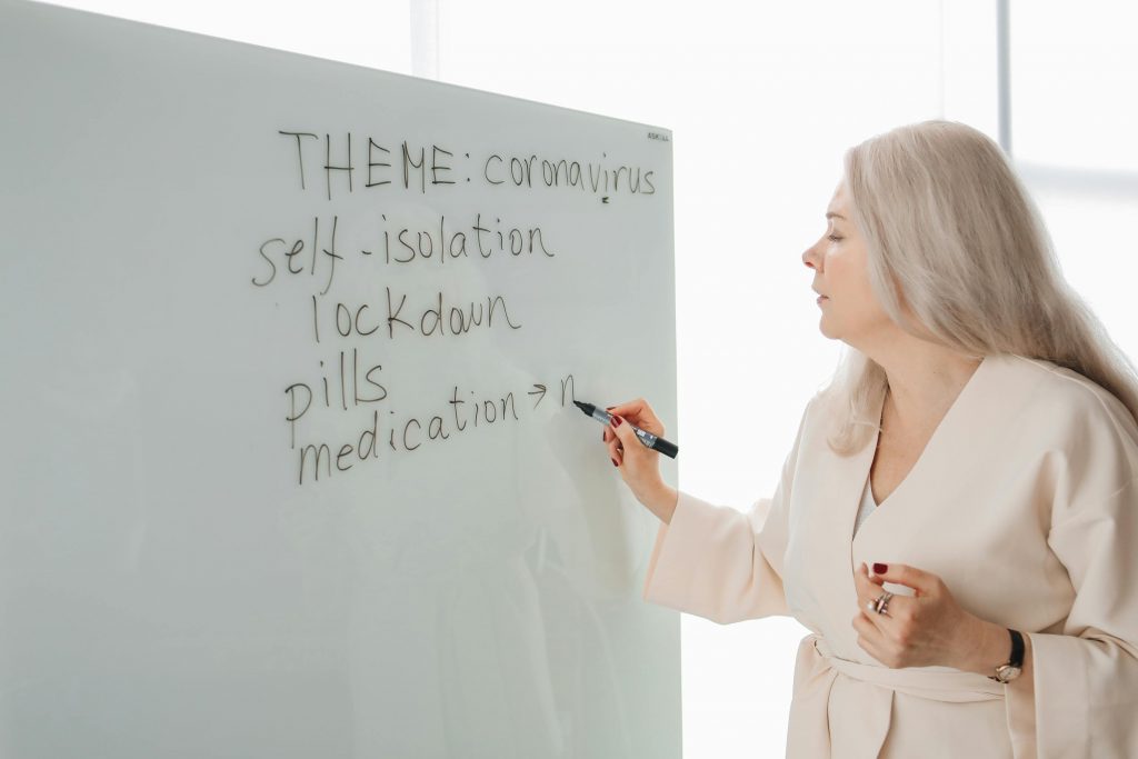 A woman instructs about coronavirus theme on a whiteboard, focusing on safety measures.